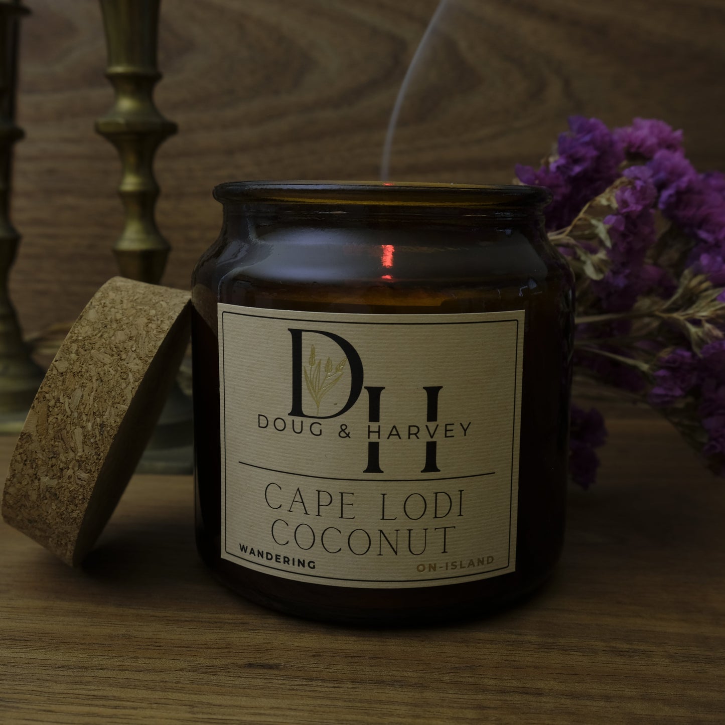 Candle jar labeled 'Doug & Harvey Cape Lodi Coconut' on a wooden surface with purple flowers and gold candlesticks in the background.
