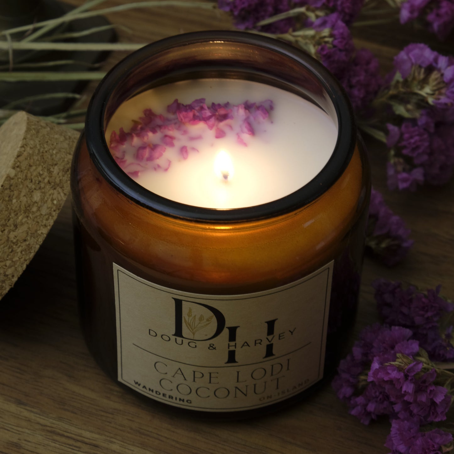 Candle in an amber jar with a label, surrounded by purple flowers on a wooden surface