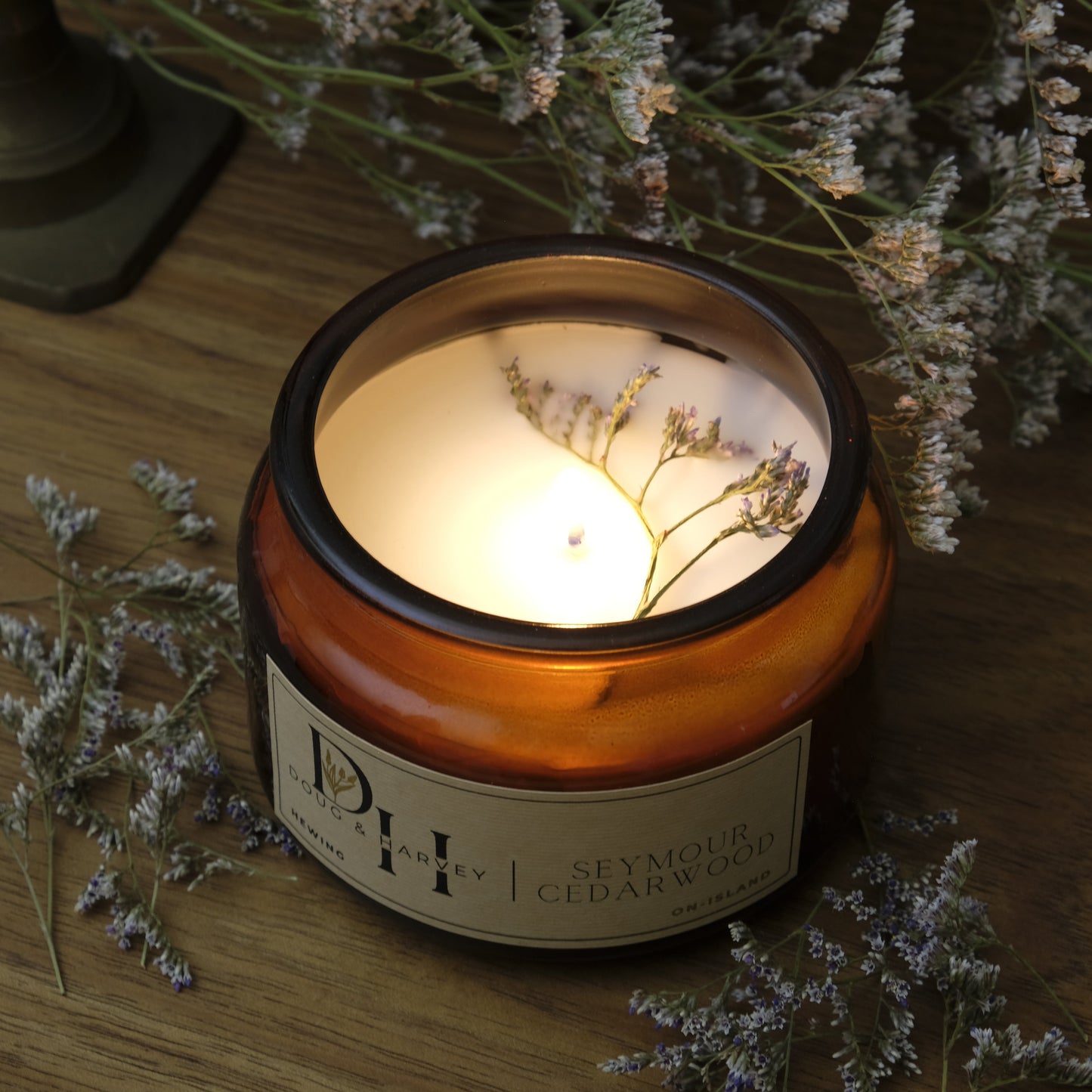 Candle in a decorative container with a label, surrounded by dried flowers on a wooden surface