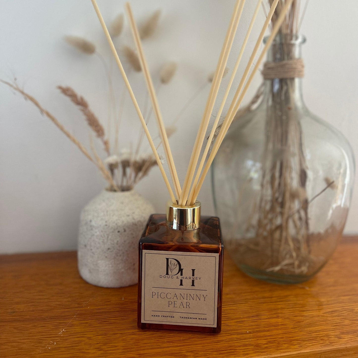 A reed diffuser with a clear glass bottle and beige label, accompanied by natural rattan reeds, placed on a wooden surface with dried plants in the background.