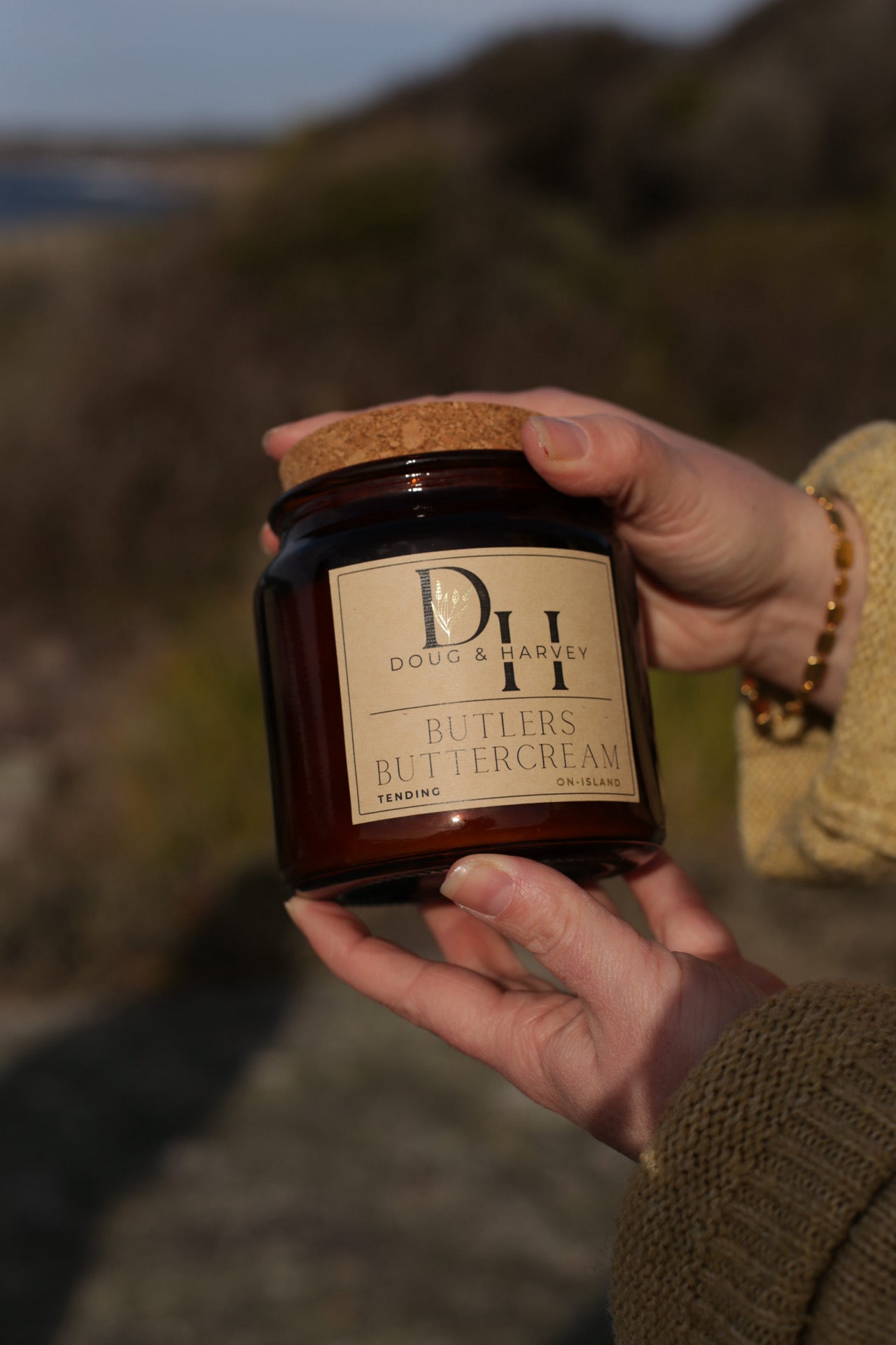 Hands holding Doug & Harvey ‘Butlers Buttercream’ candle in an amber glass jar with cork lid, photographed outdoors in natural light with blurred coastal background.