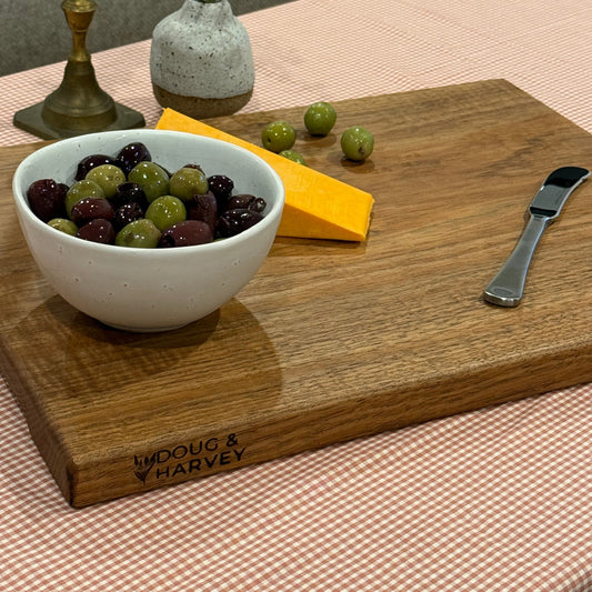 Handcrafted Tasmanian blackwood chopping board by Doug & Harvey, styled with a bowl of mixed olives, cheddar cheese, and a cheese knife on a gingham tablecloth. Features engraved Doug & Harvey branding, perfect for serving and entertaining.
