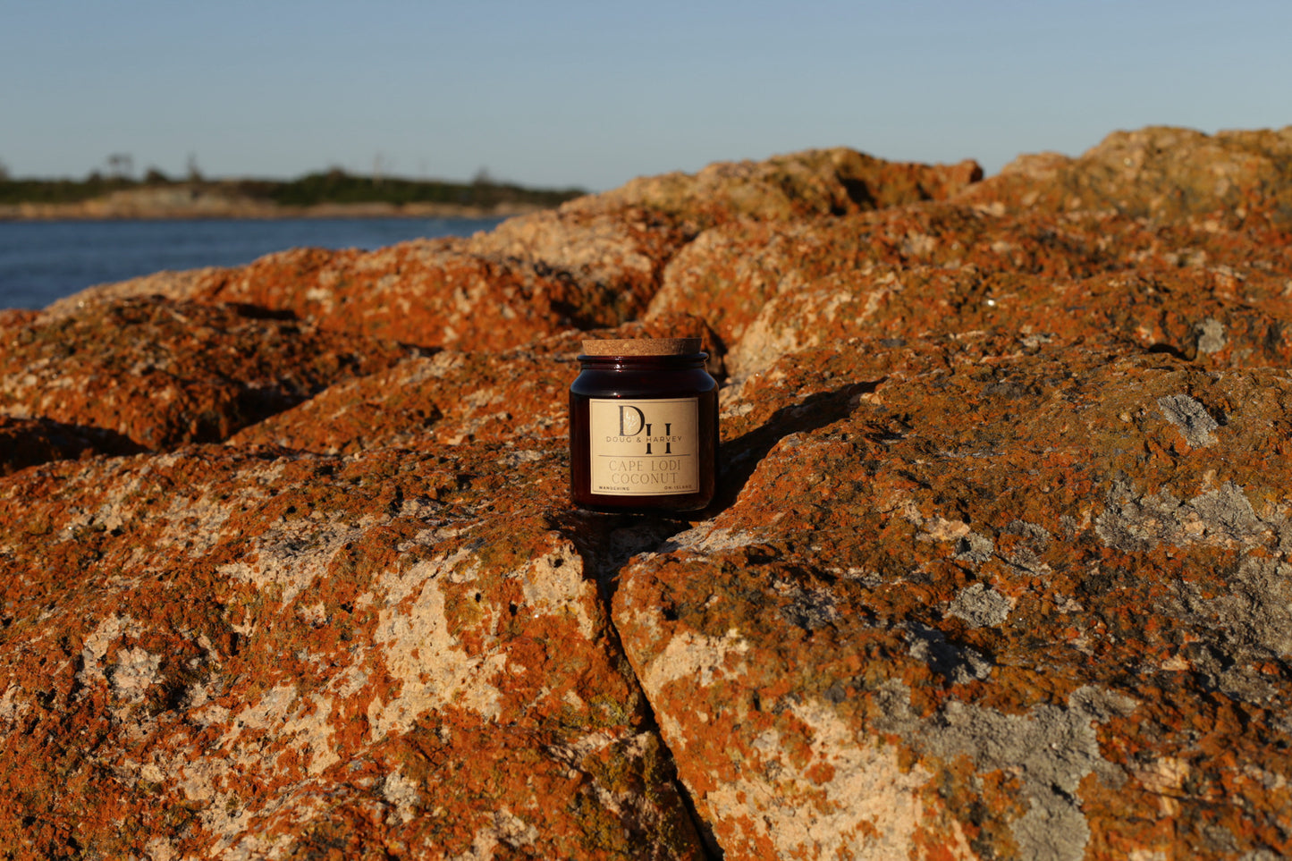 Doug & Harvey ‘Cape Lodi Coconut’ candle in an amber glass jar with cork lid, photographed outdoors on rugged orange Tasmanian rocks with the ocean and horizon in the background.