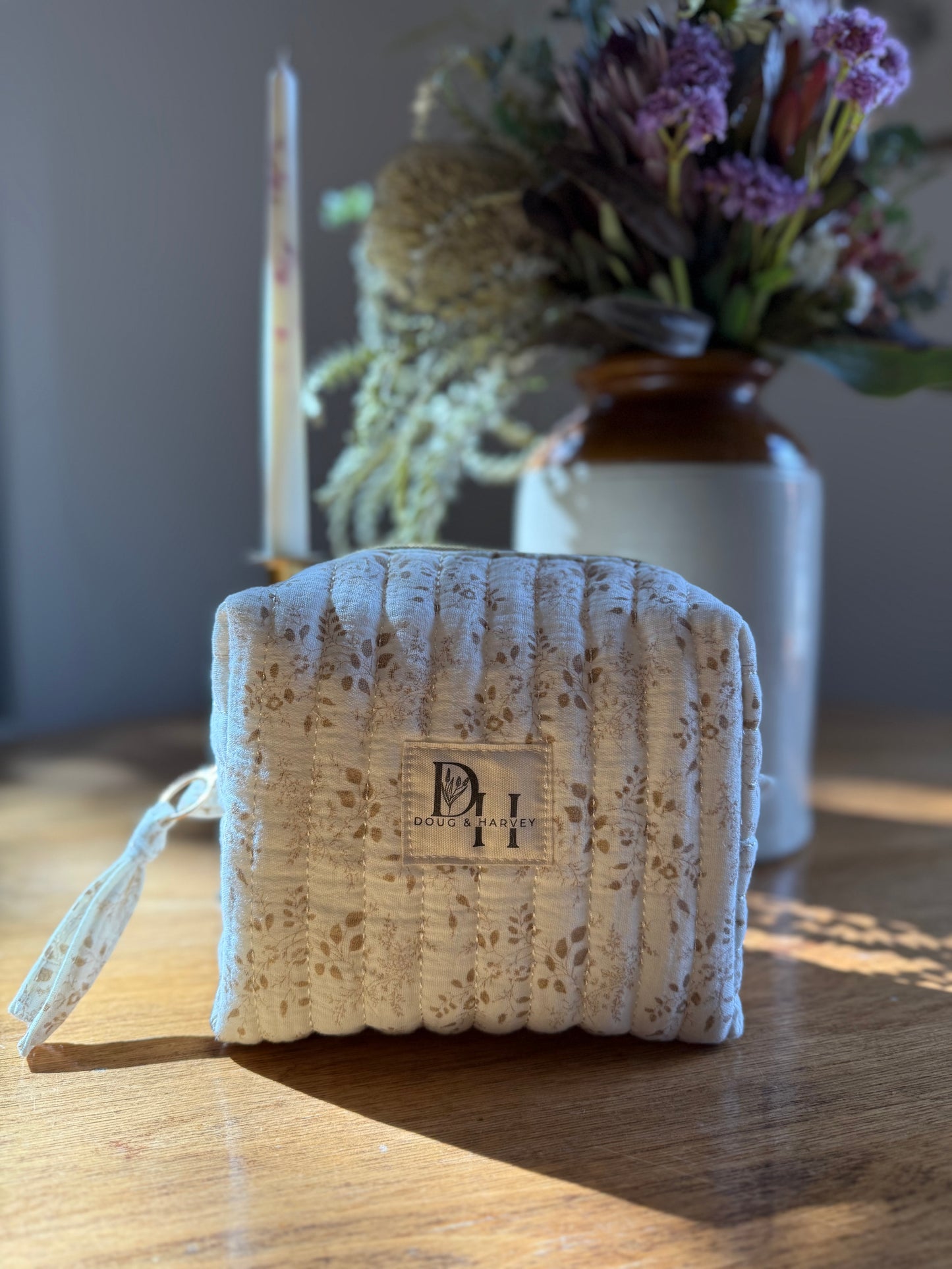Doug & Harvey quilted toiletry bag in cream floral fabric, photographed indoors on a wooden table with soft natural light, floral arrangement and candle in the background.