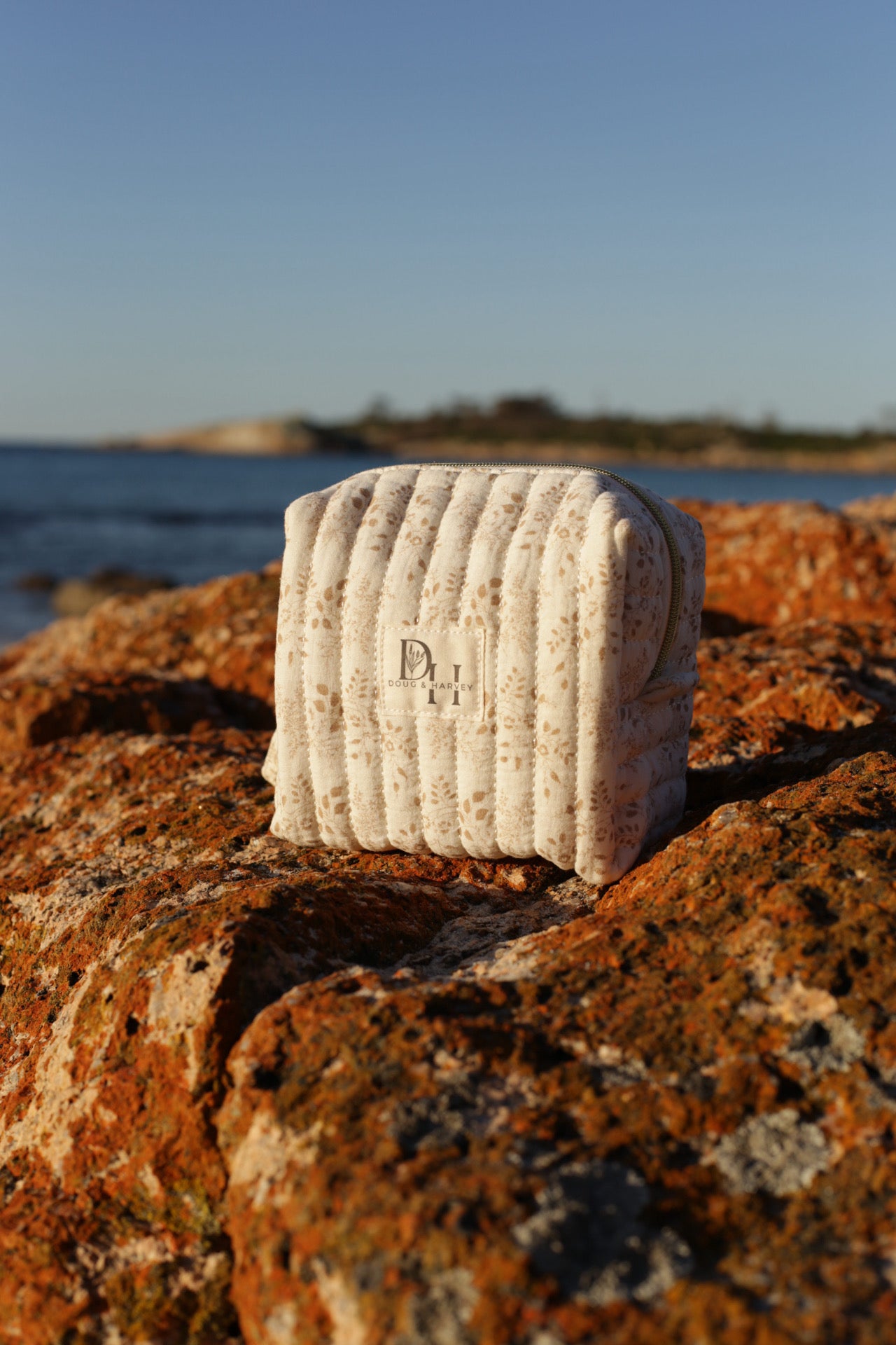 Doug & Harvey quilted toiletry bag in cream floral fabric, styled outdoors on sunlit Tasmanian rocks with ocean and coastline in the background.