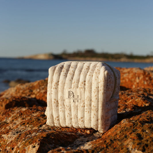 Doug & Harvey quilted toiletry bag in cream floral fabric, styled outdoors on sunlit Tasmanian rocks with ocean and coastline in the background.