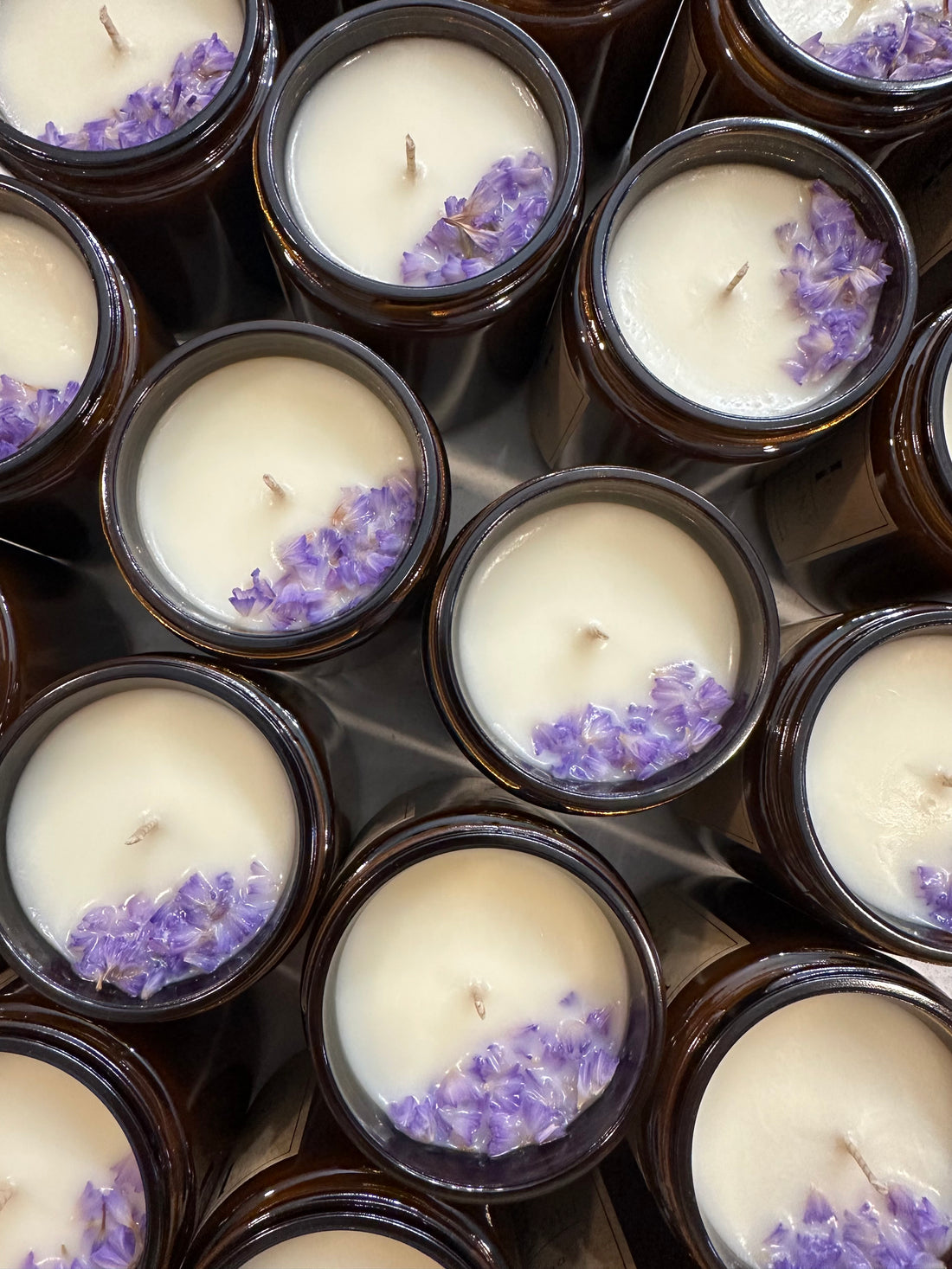 Hand-poured soy candles in amber jars, topped with delicate purple flower petals, displayed in a close-up view. The candles are arranged neatly, showcasing the natural beauty of the lavender accents against the creamy white wax.