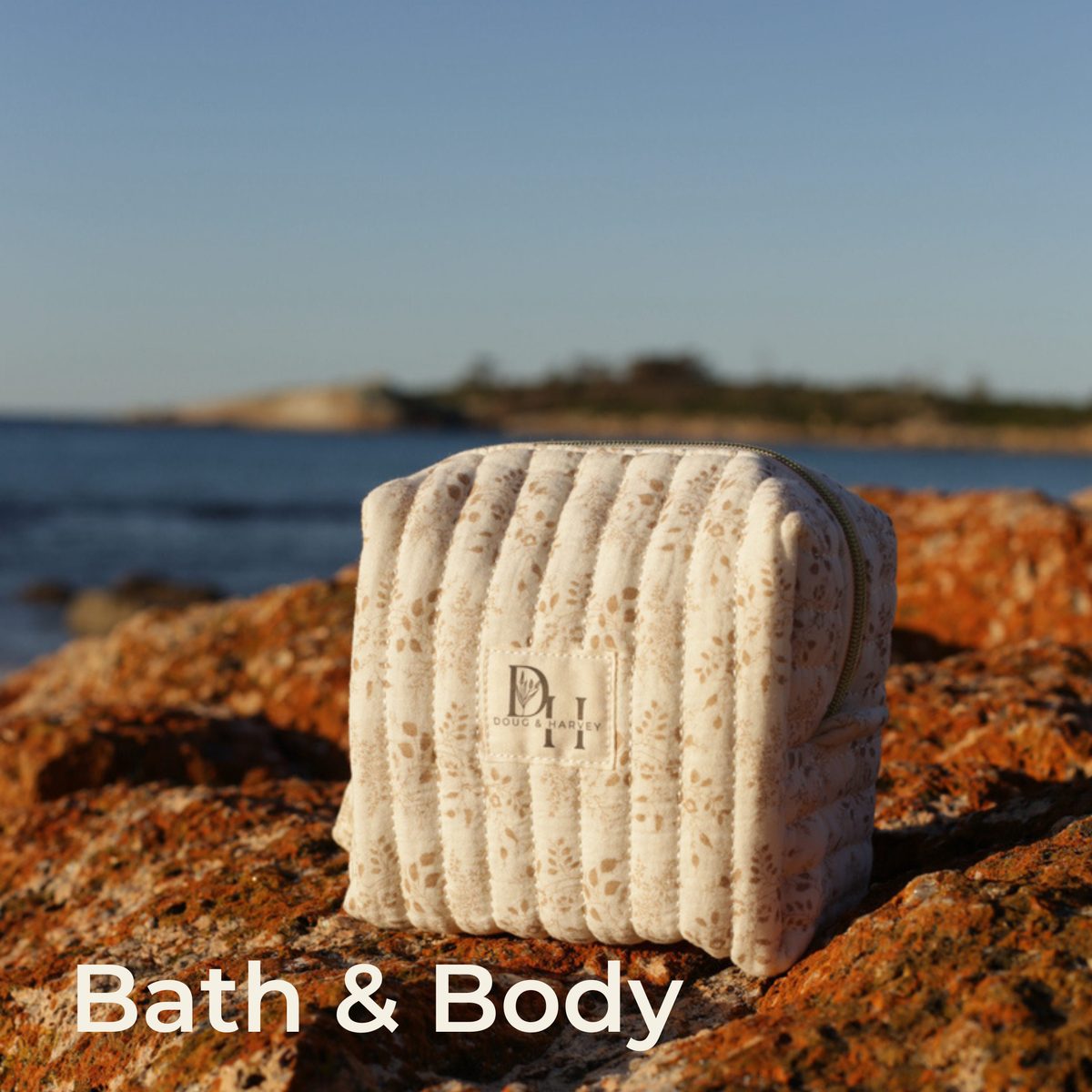 Bath & Body collection: Doug & Harvey cream quilted floral cosmetic bag on orange coastal rocks beside the sea.