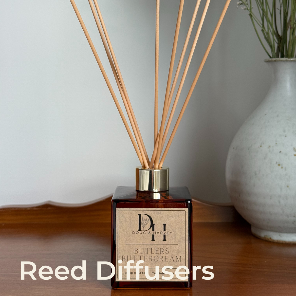 Reed Diffusers collection: Doug & Harvey Butlers Buttercream diffuser in amber glass with a gold collar, on timber beside a ceramic vase.