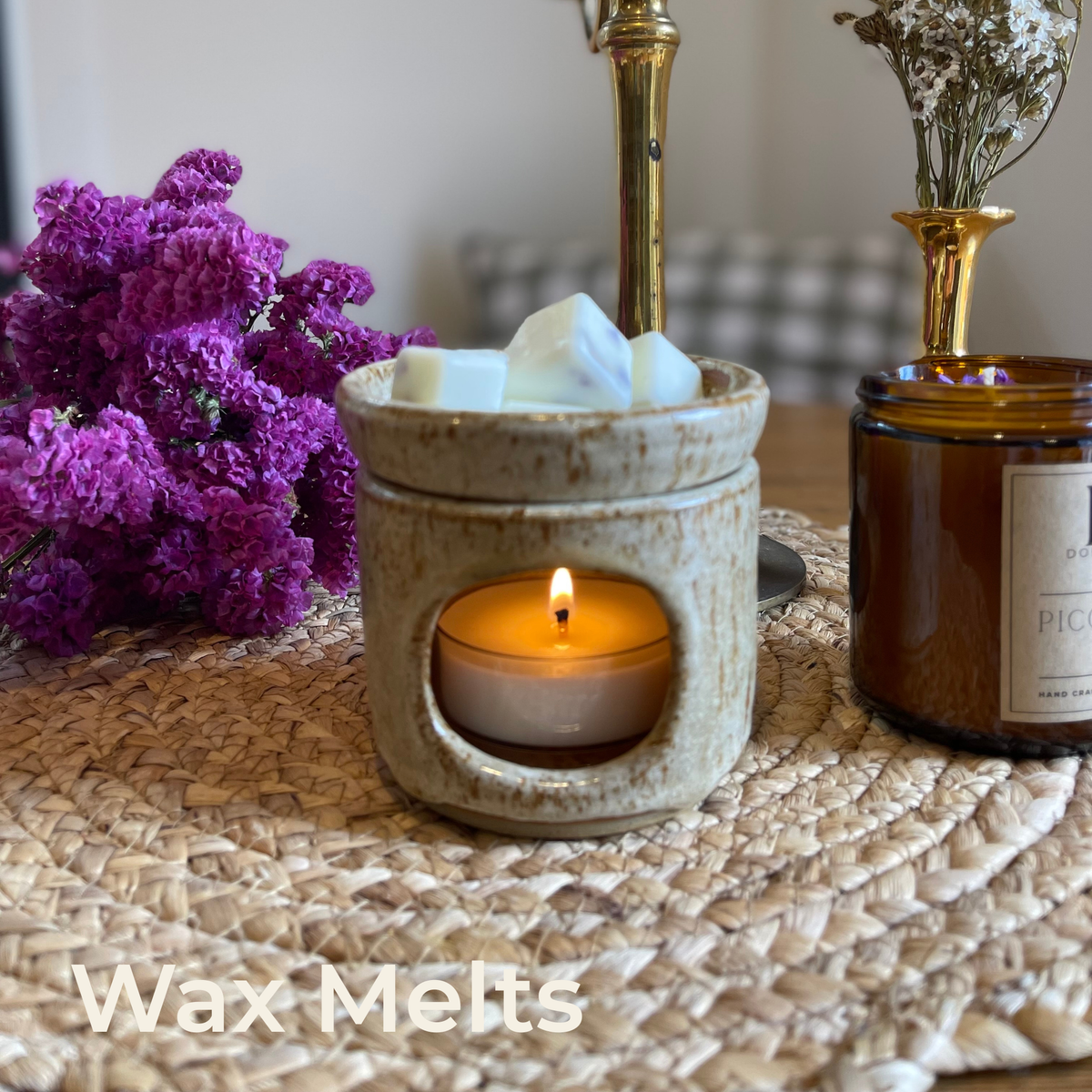 Wax Melts collection: white wax melts in a speckled ceramic burner with a lit tealight, beside purple flowers on a woven mat.
