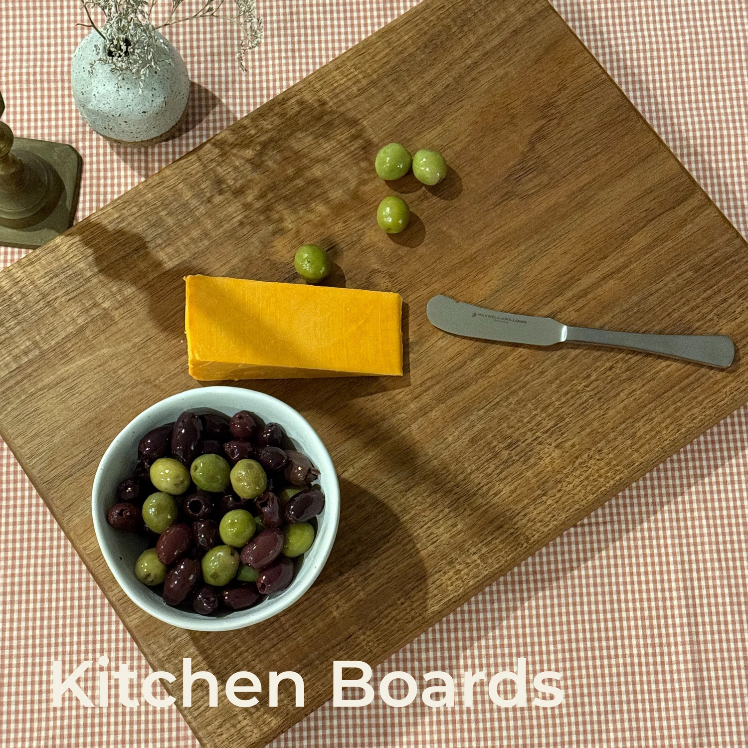 Kitchen Boards collection: a timber serving board with cheese, olives and a butter knife on a gingham tablecloth.