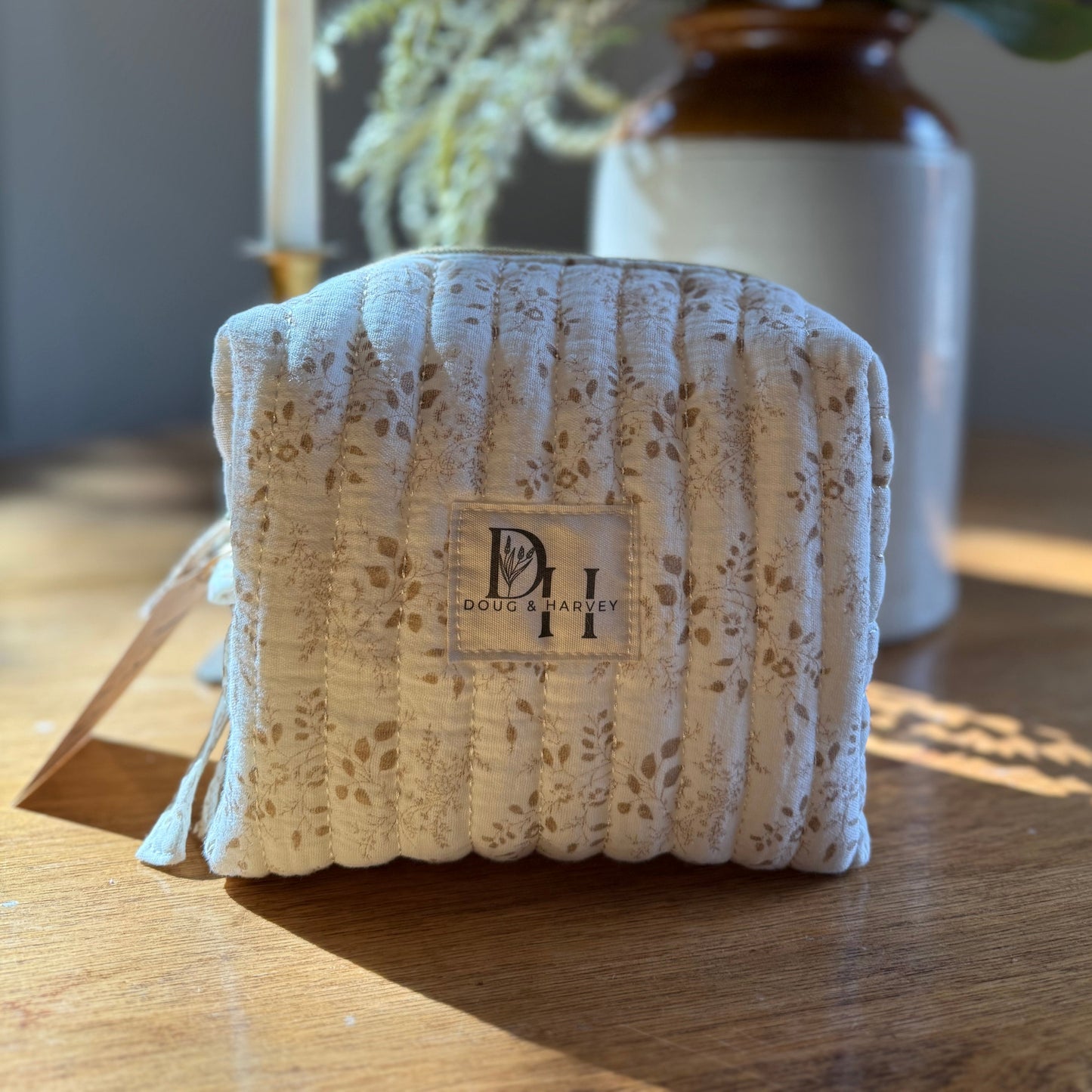 A beige quilted toiletry bag with a floral pattern, placed on a table with a vase of flowers in the background. Tasmania, Australia, Bicheno Doug and Havey 