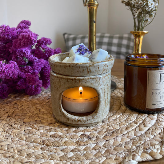 A wax melt in a beige holder with a lit candle below it, placed on a table with a woven placemat, next to a vase of purple flowers and a book.
