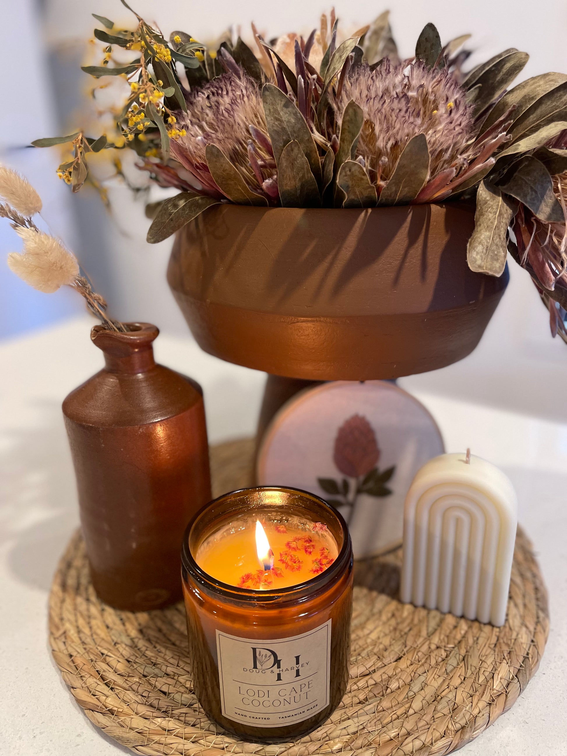 Image of lit Cape Lodi Coconut candle with pink dried native Australian flowers floating at the top of the candle set on a rattan round mat with a rainbow pillar candle, hand embroidered protea hoop, brown vase with bunny tails and ceramic vase with dried proteas and golden wattle