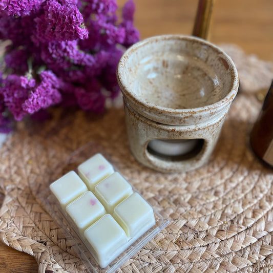 Wax melts in white and yellow colors are presented alongside a ceramic warmer on a woven mat, with purple flowers in the background.