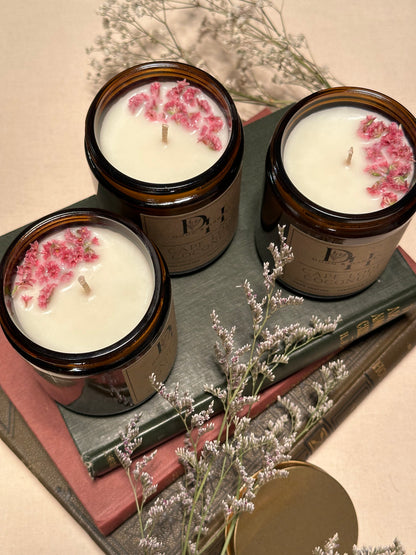 Three Doug and Harvey Cape Lodi Coconut candles with amber glass jars, brown labels, and gold tin lids are arranged on vintage books. Each candle is adorned with dried flowers, creating a charming, elegant display that highlights their rustic and sophisticated appeal.