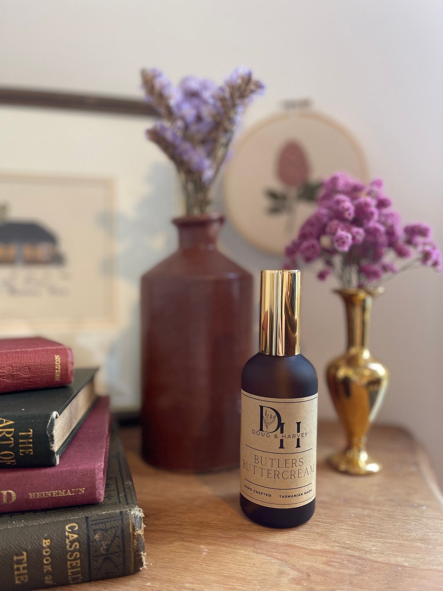 A product shot showcasing the indulgent Doug & Harvey Butlers Buttercream room spray. The brown glass bottle with a gold atomizer is elegantly displayed amidst books, a brass vase with purple flowers, and other decorative accents, emphasizing the product's sophisticated appeal and the brand's association with refined living