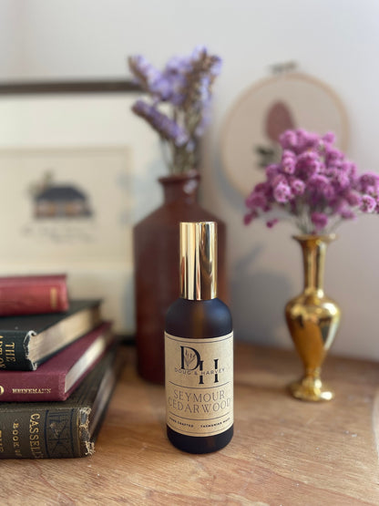 A product shot of a brown glass spray bottle with a gold atomizer, labeled "Seymour Cedarwood" and featuring the Doug & Harvey logo. The bottle is surrounded by books, a brass vase with purple flowers, and another decorative vase. This image highlights the product's aesthetic appeal and the brand's association with refined living