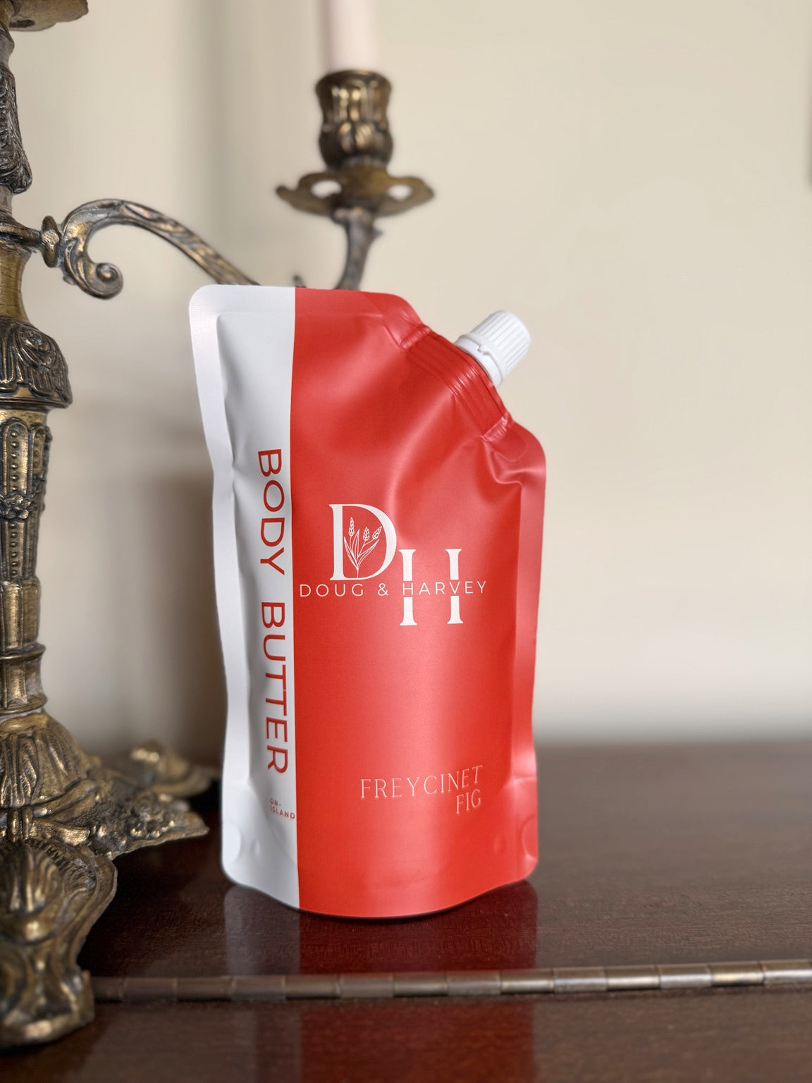 Doug & Harvey Freycinet Fig Body Butter in a coral-and-cream pouch on a dark timber piano beside an ornate gold candelabra.