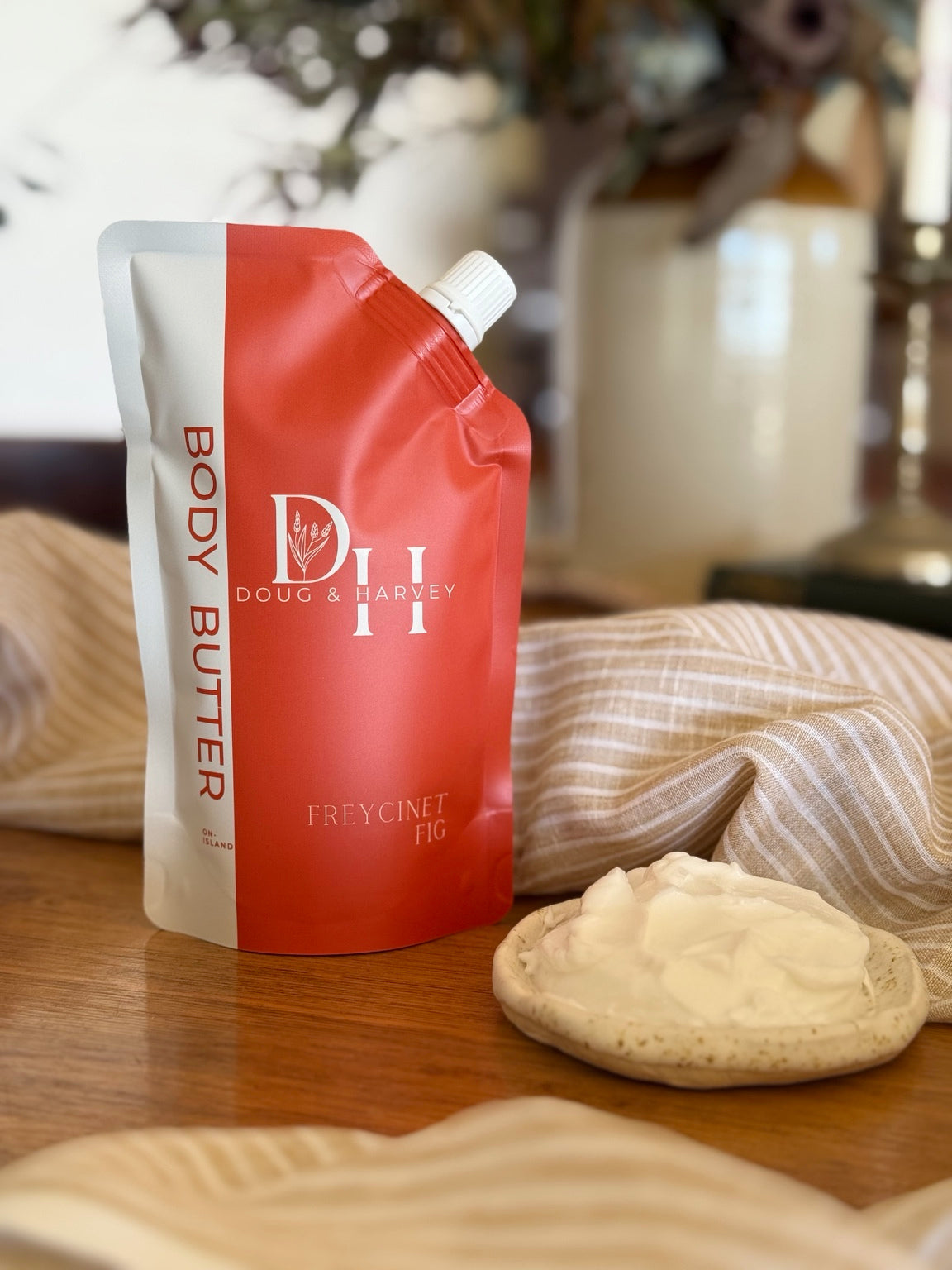 Doug & Harvey Freycinet Fig Body Butter in a coral-and-cream pouch beside a dish of body butter, styled on timber with striped linen.