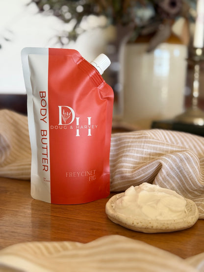 Doug & Harvey Freycinet Fig Body Butter in a coral-and-cream pouch beside a dish of body butter, styled on timber with striped linen.