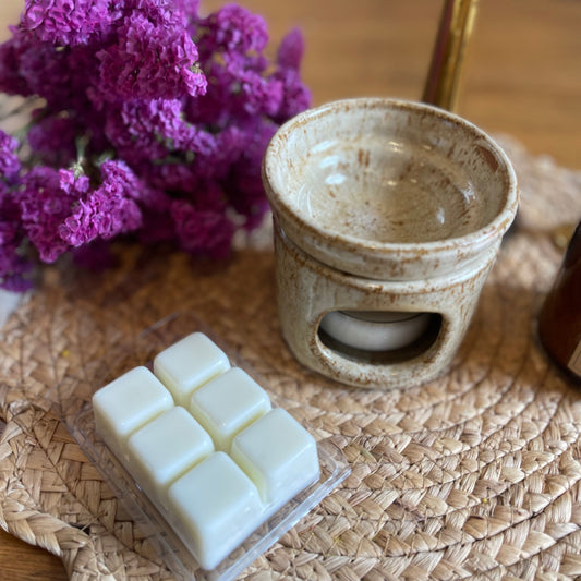 A beige wax melt container with a lid, placed on a rattan mat. The container is adorned with a small bouquet of purple flowers, and there are several off-white wax melts visible in an open square tray beside it.