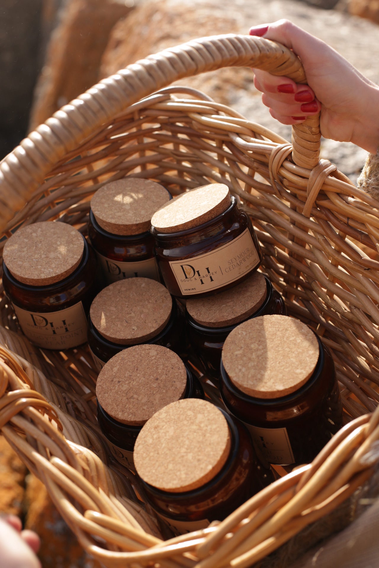 Basket filled with Doug & Harvey amber glass candles with cork lids, carried outdoors in natural sunlight, showing multiple scents including Seymour Cedarwood and Peggy’s Point Pear.