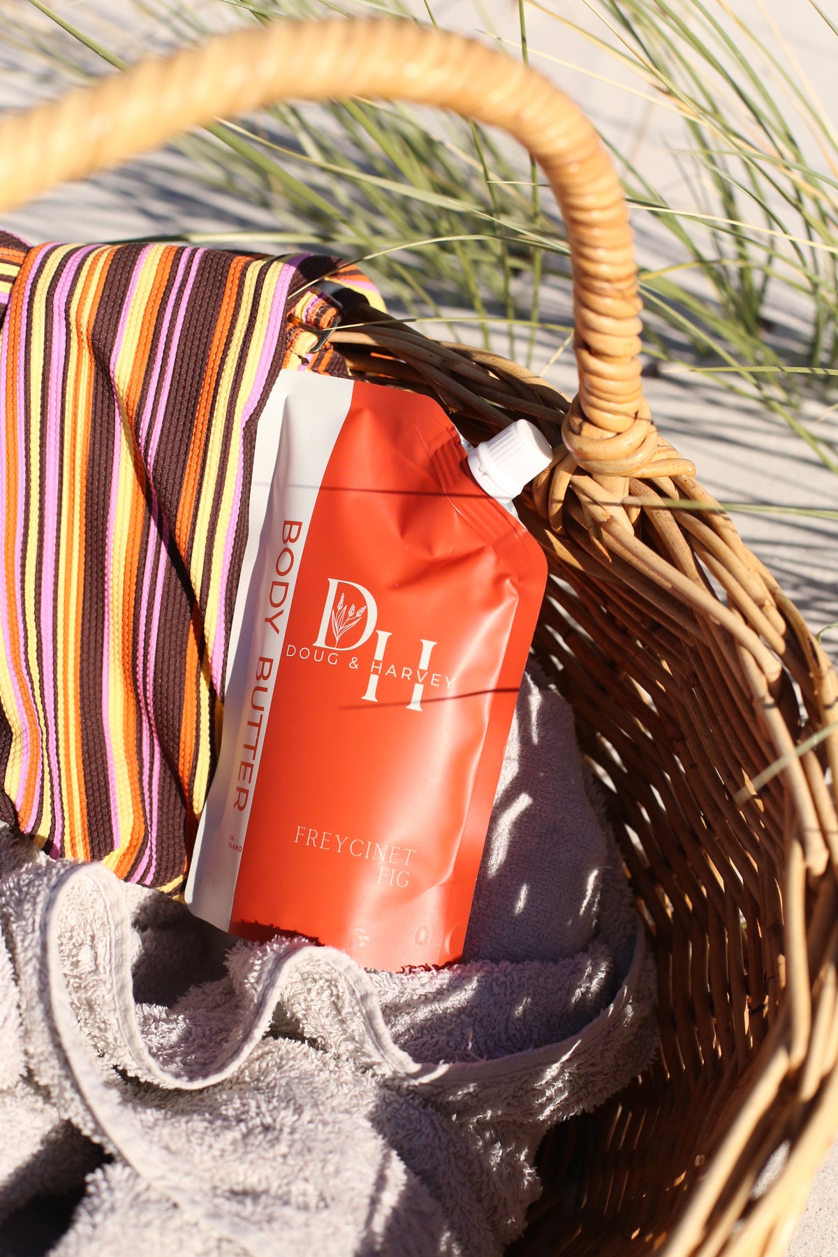 Doug & Harvey Freycinet Fig body butter pouch nestled in a wicker beach basket beside a grey towel and striped fabric.
