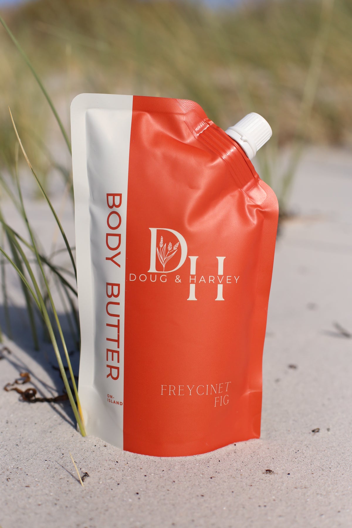 Doug & Harvey Freycinet Fig body butter in an orange and white pouch standing on beach sand beside coastal grasses.