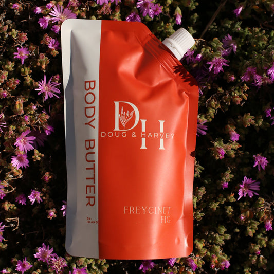 Doug & Harvey Freycinet Fig body butter in an orange and white pouch nestled among purple flowers. 