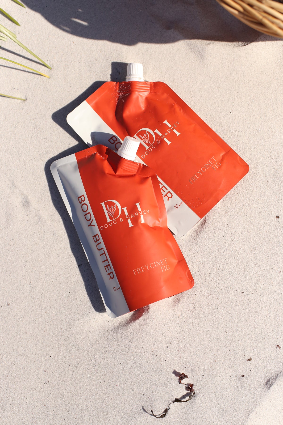 Two orange and white pouches of Doug & Harvey Freycinet Fig body butter lying on sunlit beach sand.
