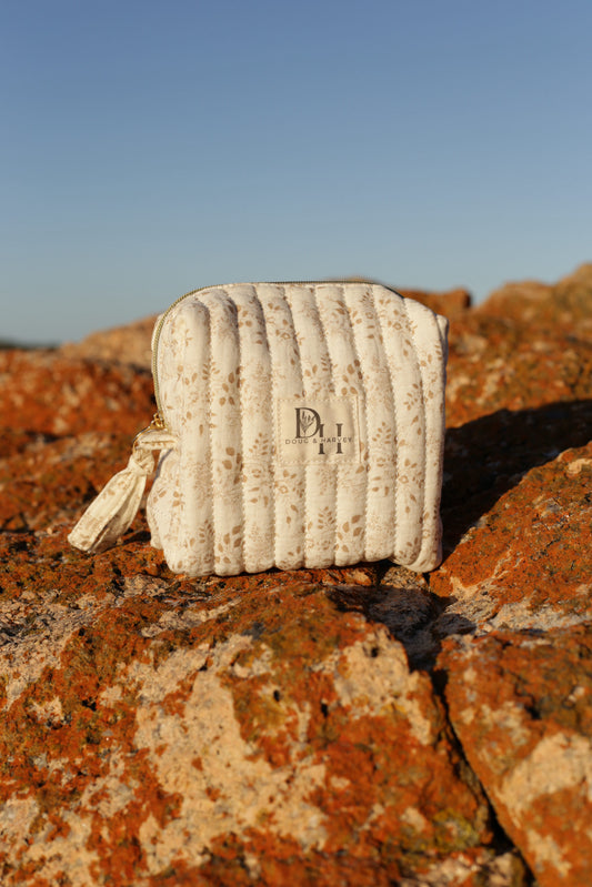 Doug & Harvey quilted toiletry bag in cream floral fabric with zip closure and side handle, photographed on sunlit Tasmanian rocks with clear blue sky.