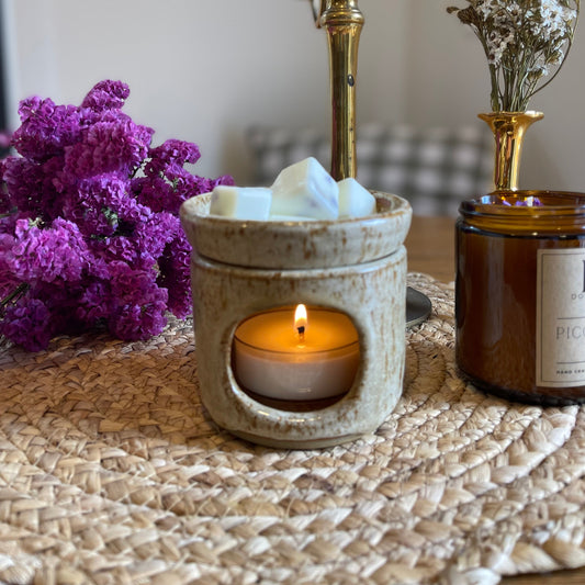 A wax melt from the brand Doug & Harvey is shown in a white ceramic warmer, with a lit candle below it. In the background, there are decorative items and a vase with purple flowers.