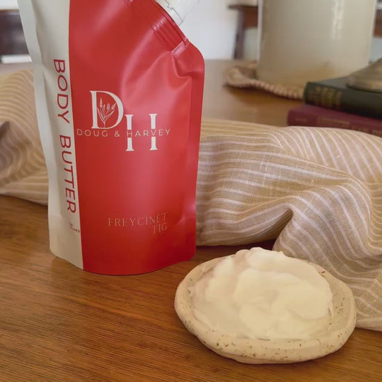 Doug & Harvey Freycinet Fig Body Butter video featuring the coral-and-cream pouch beside a dish of body butter, styled with striped linen on timber.