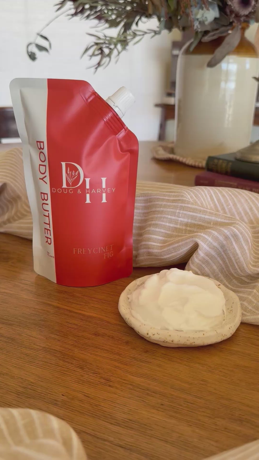 Doug & Harvey Freycinet Fig Body Butter video featuring the coral-and-cream pouch beside a dish of body butter, styled with striped linen on timber.