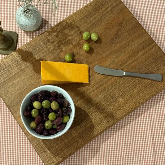 Handcrafted Tasmanian blackwood chopping board styled with cheddar cheese, green and kalamata olives, and a butter knife on a gingham tablecloth. A versatile and timeless serving board, perfect for cheese platters and entertaining.